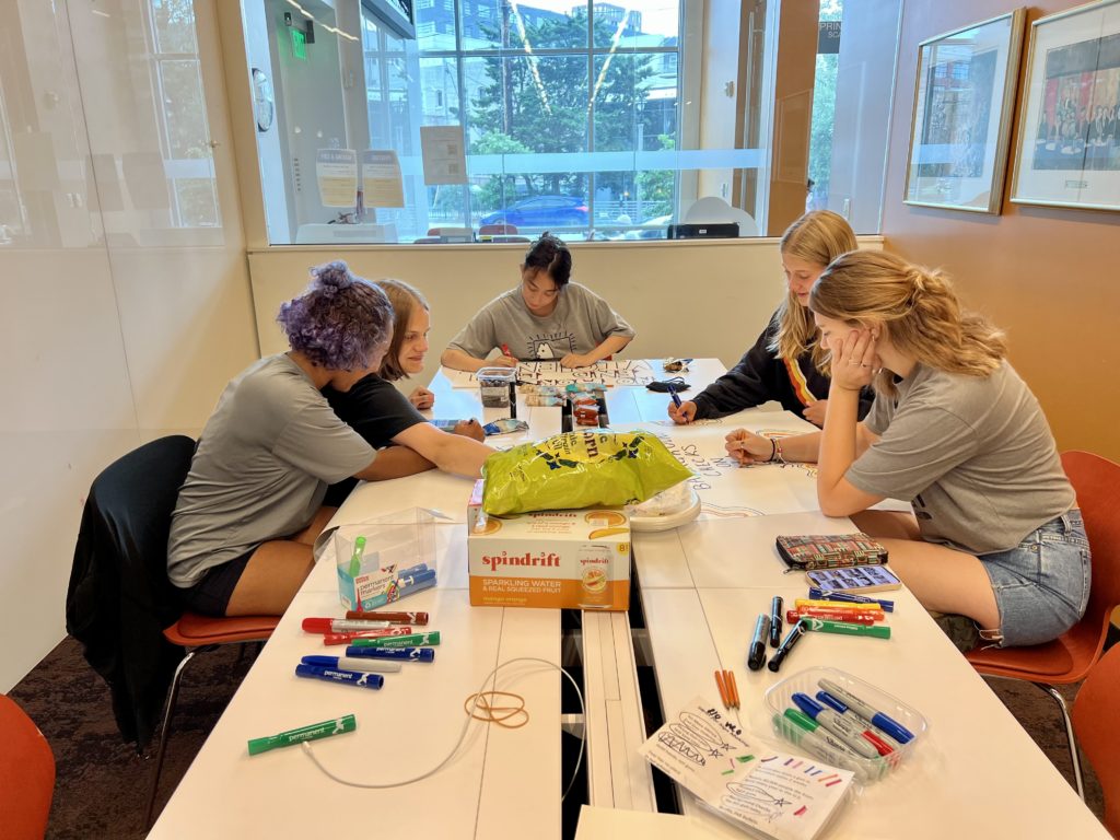 Five young women sitting around a table writing on poster board. 