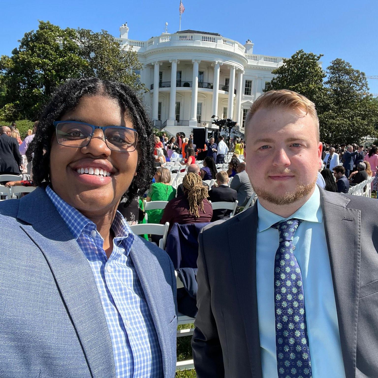 Two WAVE employees are in the foreground smiling at the camera. They are wearing suits. In the background are many people seated, and in the far background is the White House. WAVE's activities and achievements earned invitations by the U.S. President.